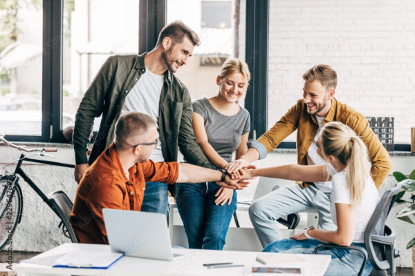 group of smiling young entrepreneurs making team gesture while working on startup together at office