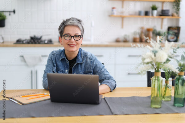 Senior woman at workplace in home interior, smiling and typing on laptop.